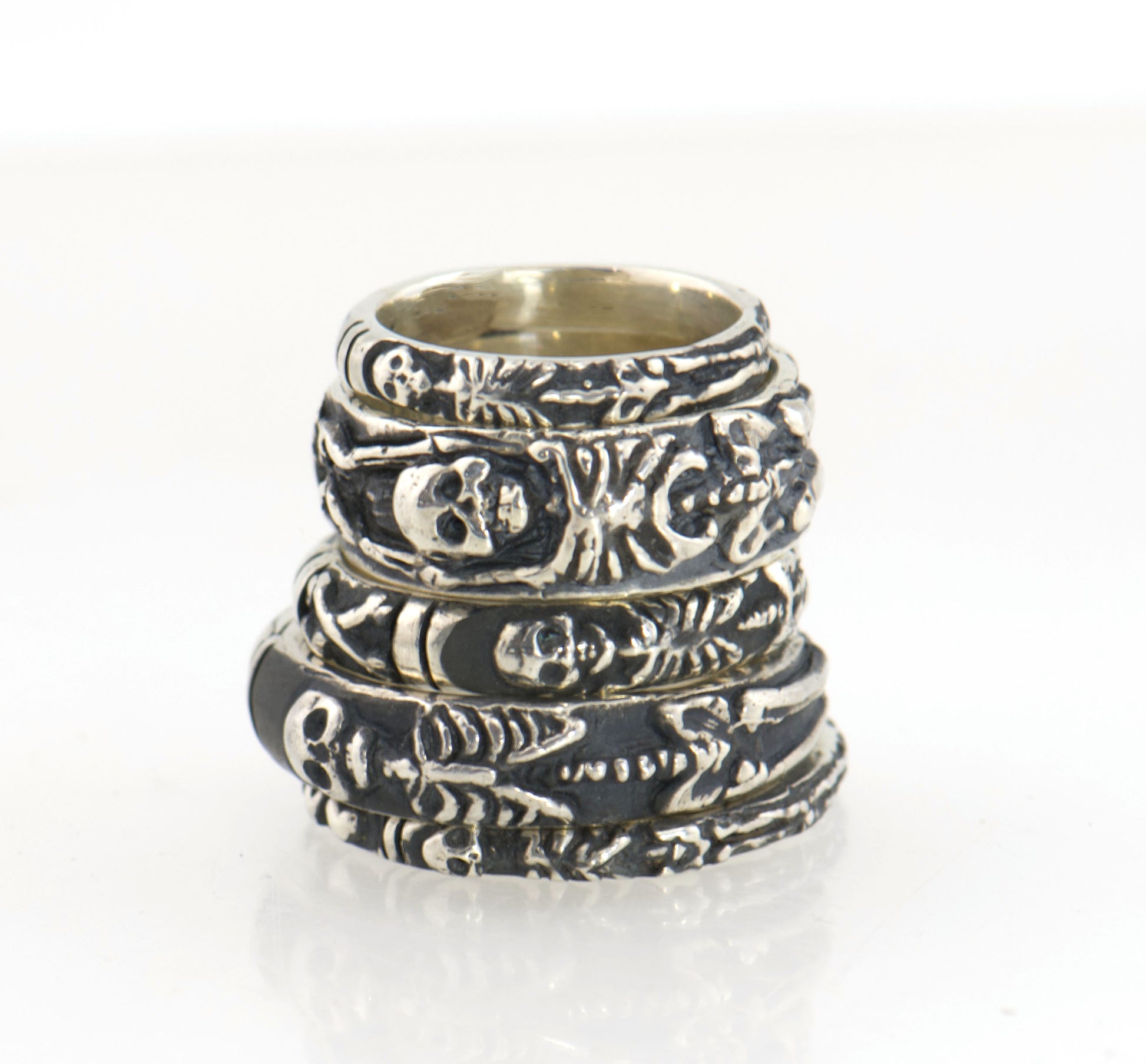 A stacked set of 5 silver rings featuring detailed,  raised skeleton motifs, including a skull and rib cage design, with a polished interior. Sterling Silver